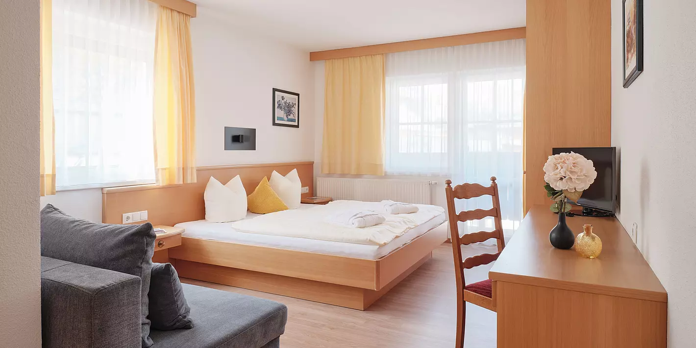 Zimmer mit Doppelbett, Schreibtisch aus Holz, grauer Couch, Holzschrank und 3 größeren Fenstern