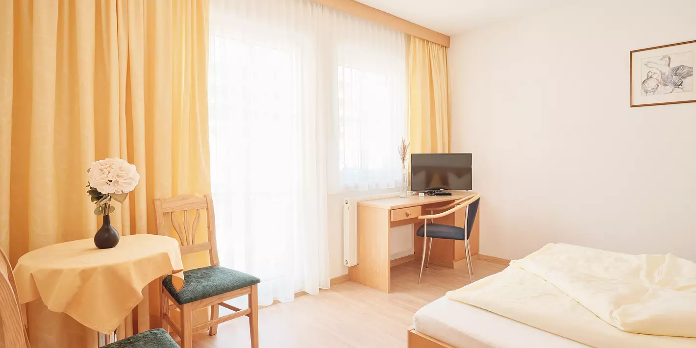 Zimmer mit kleinem Holzschreibtisch, Sitzecke, Bett und großen Fenster