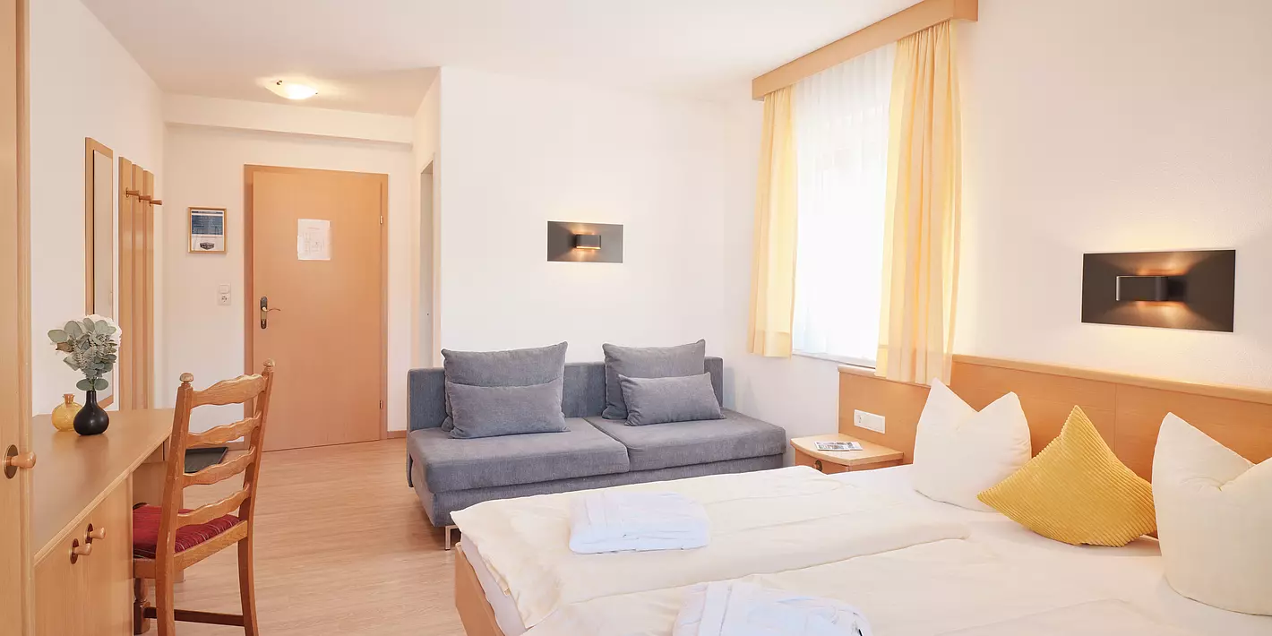 Zimmer mit Doppelbett, Holzschreibtisch, grauer Couch und Fenster